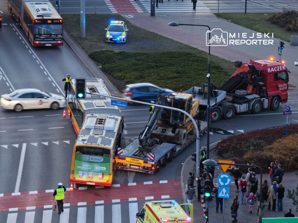 Wielki autobus komunikacji miejskiej stoi w zablokowanym ruchu, a obok niego pracuje ciężki sprzęt budowlany. Policjant kieruje ruchem, a w tle widać inne pojazdy oraz grupę przechodniów.