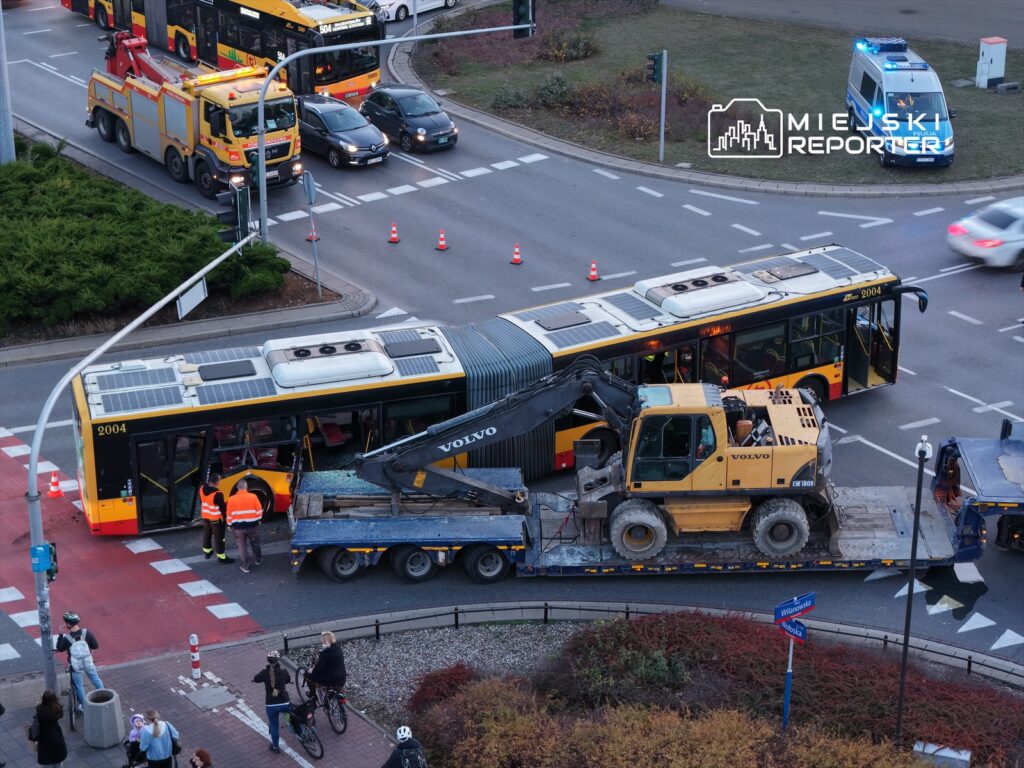 Zespół pracowników w odblaskowych kamizelkach bada uszkodzony autobus na skrzyżowaniu, obok ciężarówki z koparką.