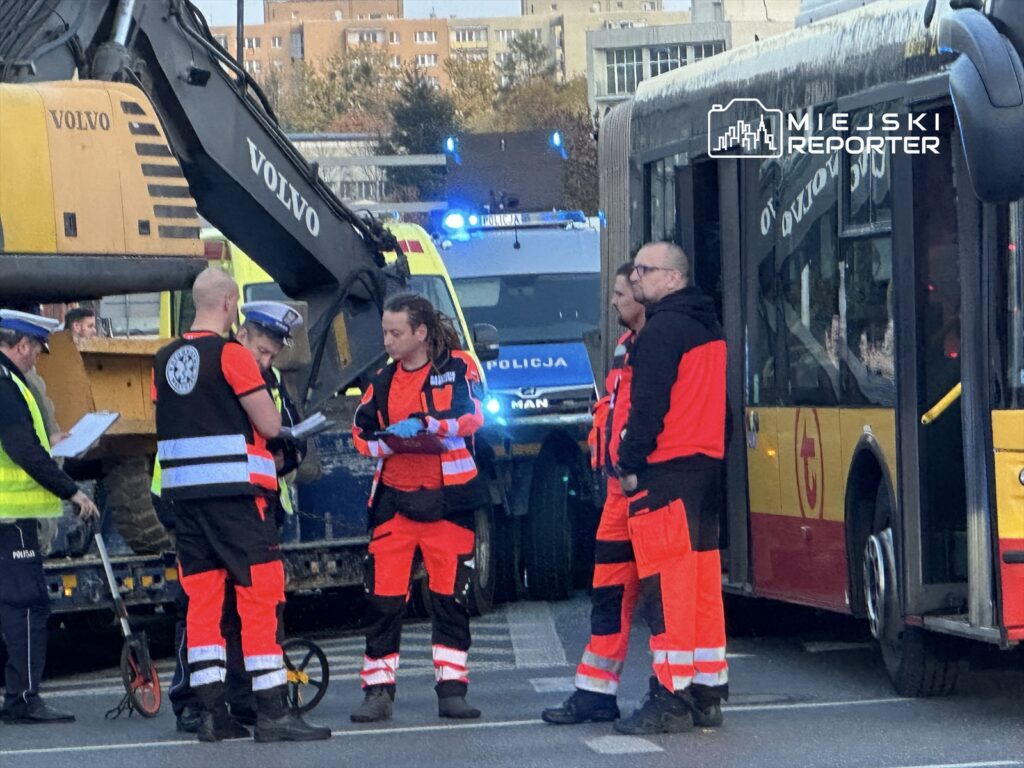 Grupa ratowników i policjantów rozmawia obok żółtego autobusu i koparki na miejskiej ulicy.