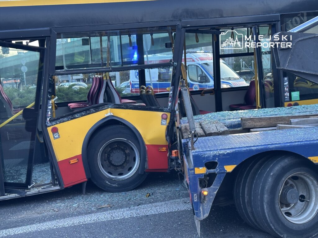Zniszczony autobus komunikacji miejskiej z wybitymi szybami stoi obok lawety na ulicy, w tle widać ambulans.