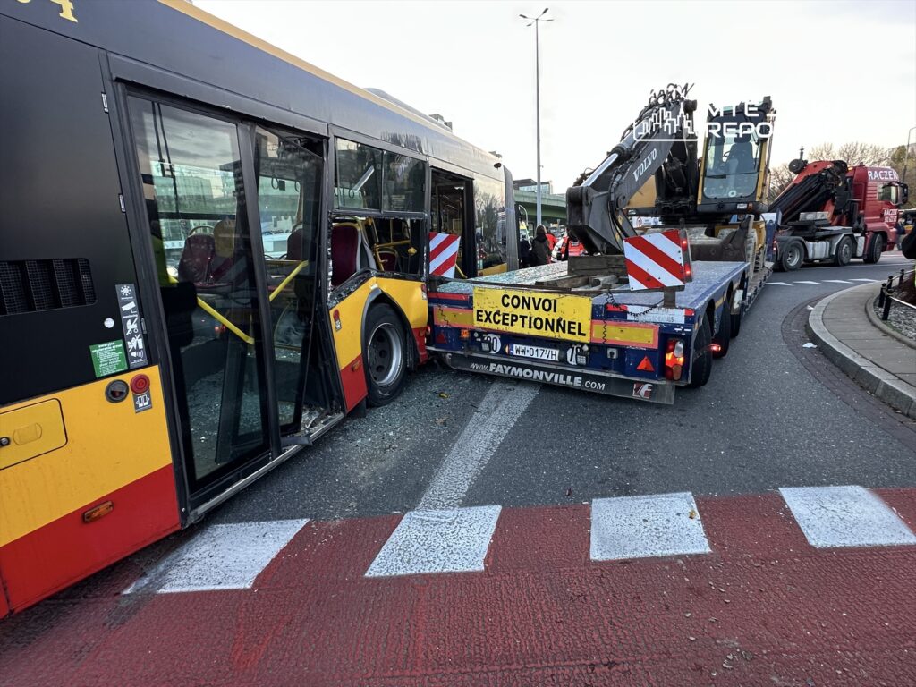 Zniszczony autobus miejski z otwartymi drzwiami stoi na skrzyżowaniu obok ciężarówki transportującej koparkę.