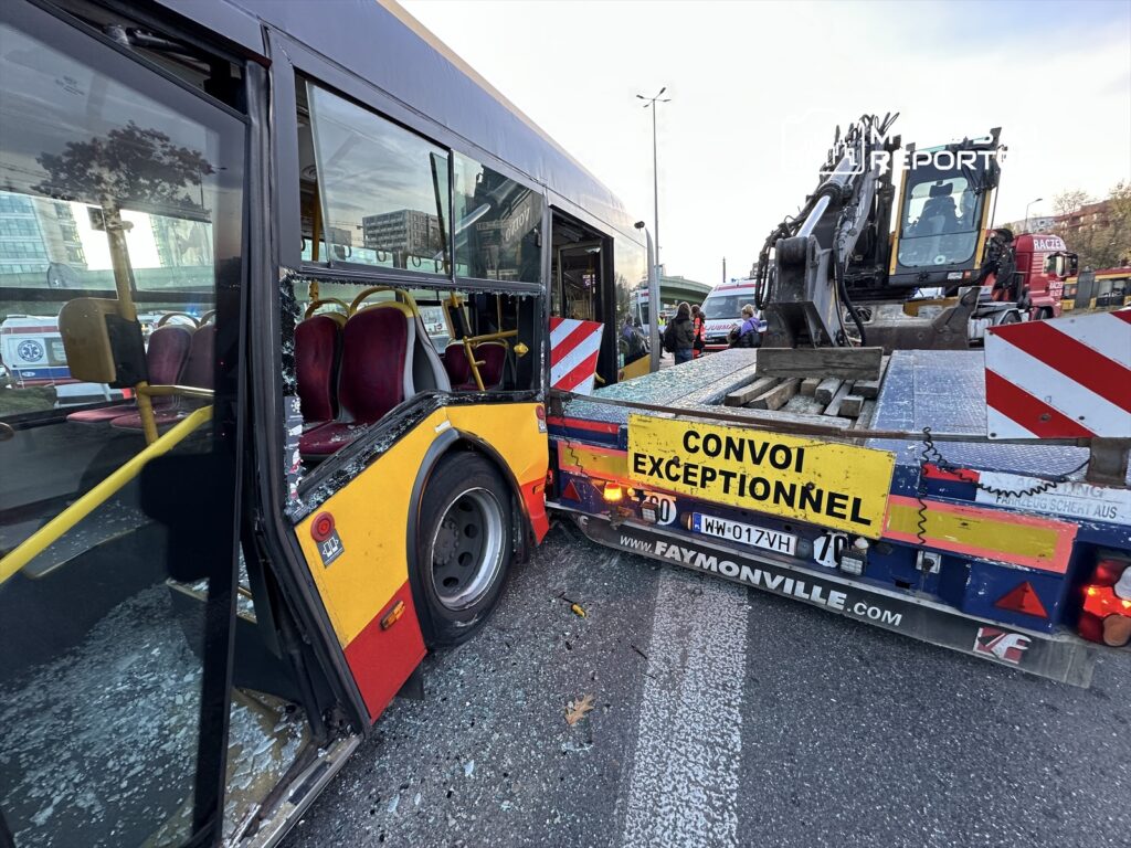 Zniszczony autobus z wybitymi szybami stoi obok ciężarówki z oznaczeniem "CONVOI EXCEPTIONNEL" na ruchliwej ulicy.