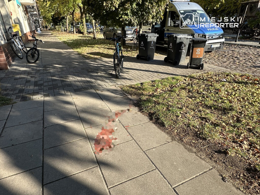 Na chodniku w miejskim otoczeniu widać dwie zaparkowane rowery oraz plamę na nawierzchni, obok stoją pojemniki na odpady.