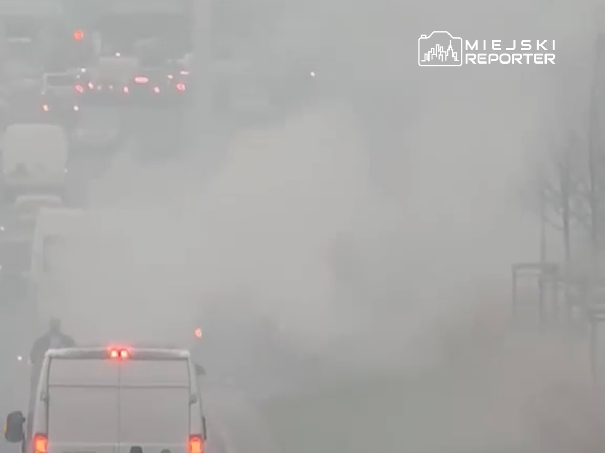 Gęsty smog ogranicza widoczność na ruchliwej drodze, gdzie stoją samochody i dostawczaki.