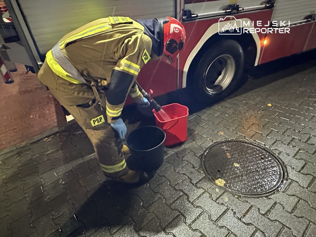 Strażak w mundurze z logo PSP nalewa wodę z czarnego wiadra do czerwonego pojemnika obok wozu strażackiego.