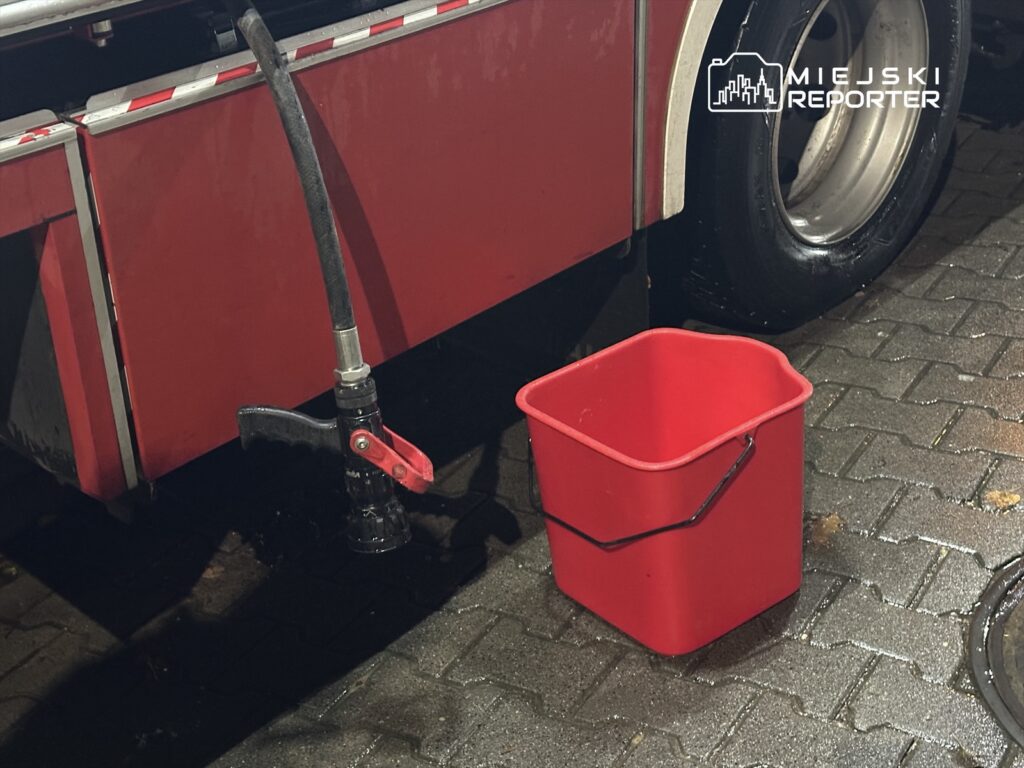 Czarna wąż z dyszą podłączony do czerwonego wozu strażackiego, obok czerwona wiadro na mokrej kostce brukowej.