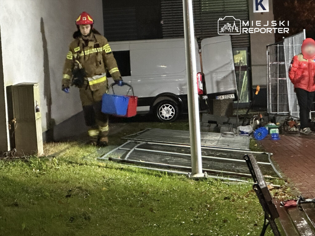 Strażak w mundurze niesie niebieską i czerwoną torbę w pobliżu budynku, obok zaparkowanego samochodu dostawczego.