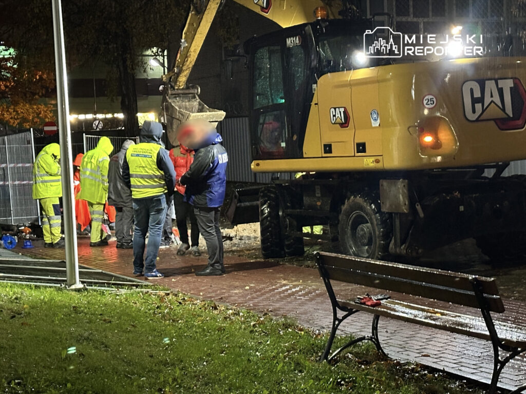 Grupa pracowników w odblaskowych kurtkach rozmawia w deszczowej scenerii obok żółtej koparki.