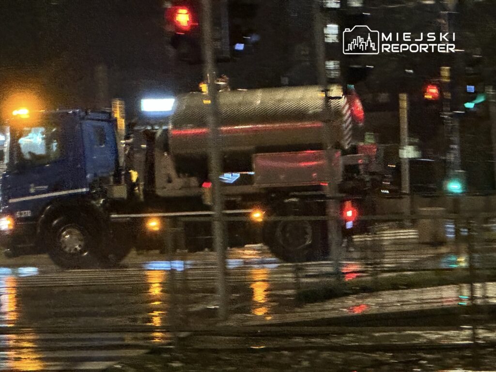 Niebieski samochód ciężarowy z cysterną przejeżdża przez mokrą ulicę w deszczową noc, mijając sygnalizację świetlną.
