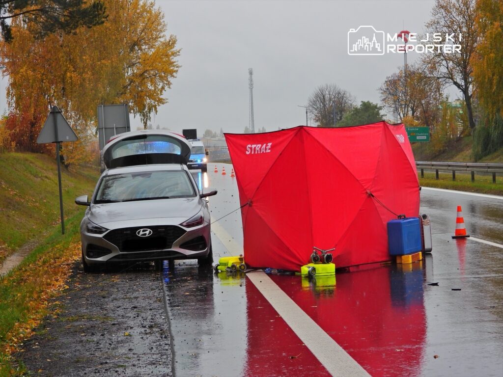 Na poboczu drogi stoi samochód osobowy z otwartym bagażnikiem, obok rozstawiony czerwony namiot straży.