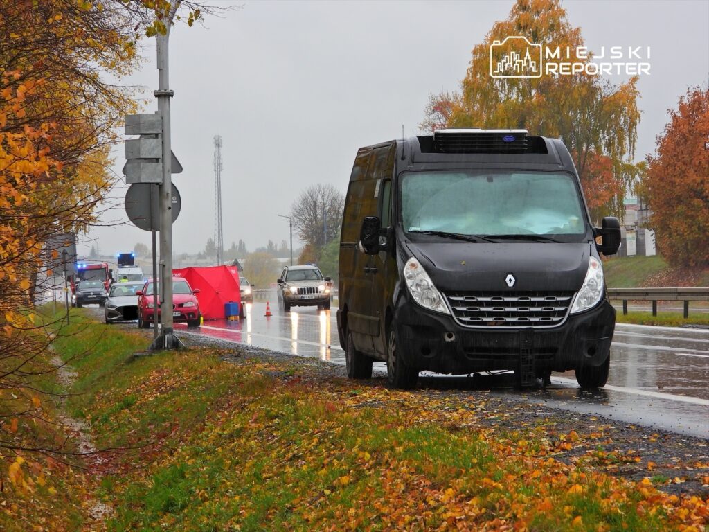 Czarny furgon zaparkowany na poboczu drogi, obok czerwony namiot i kilka samochodów, w deszczowej aurze.