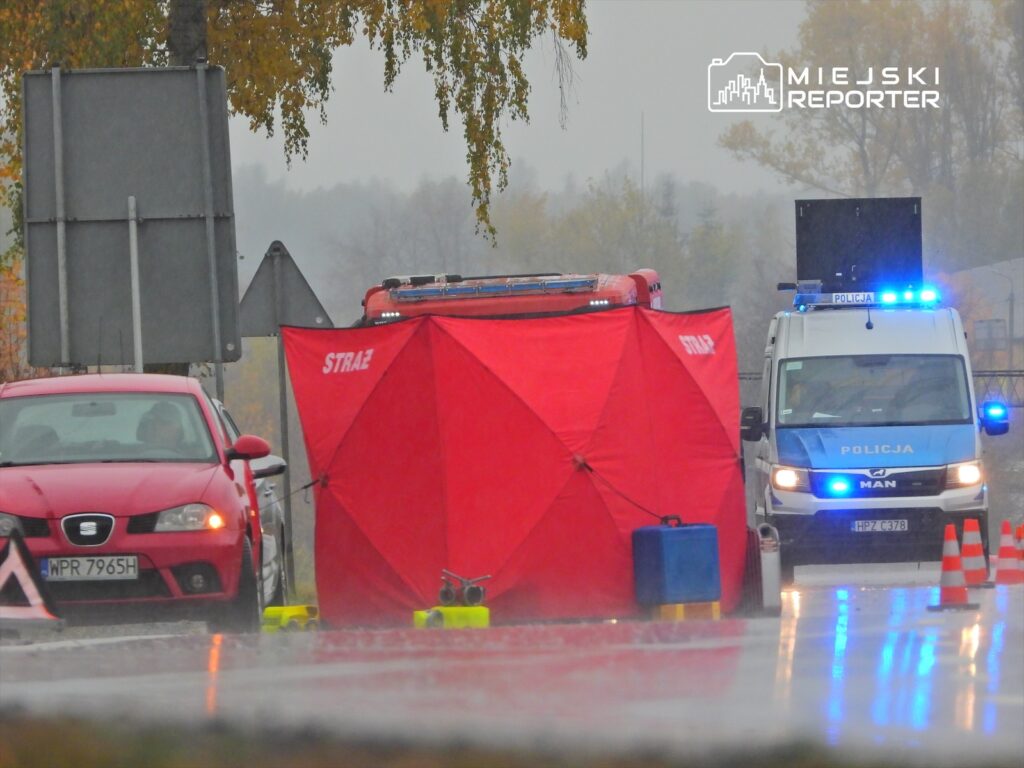 Czerwony namiot straży ustawiony obok policyjnego samochodu na mokrej drodze, w tle widoczne znaki drogowe.