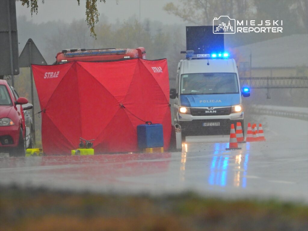 Czerwony namiot strażacki ustawiony na deszczowej drodze, obok policyjny samochód z włączonymi sygnałami świetlnymi.
