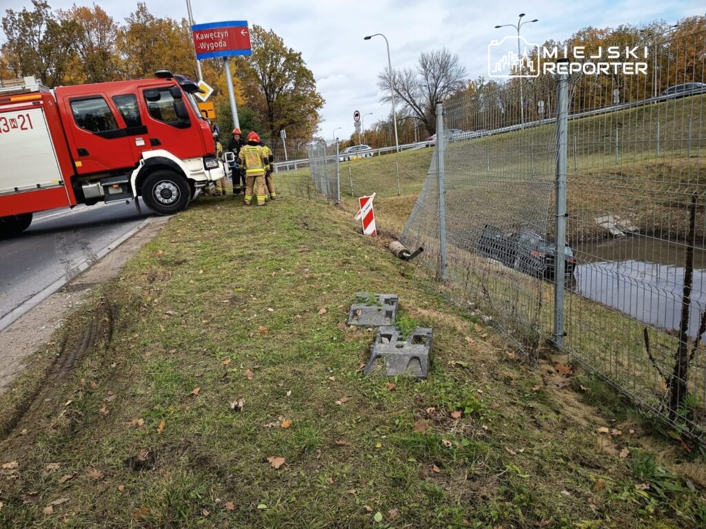 Czerwony wóz strażacki stoi na poboczu drogi, a strażacy przygotowują się do akcji ratunkowej przy rowie.