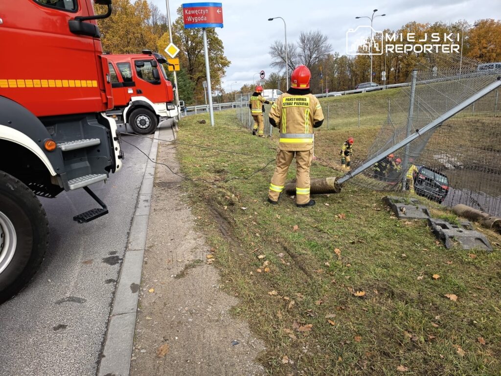 Strażacy w mundurach pracują nad usunięciem uszkodzonego ogrodzenia przy drodze, obok pojazdów strażackich.