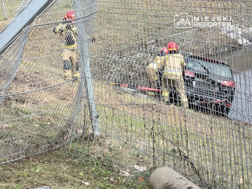 Dwaj strażacy w kaskach i mundurach wyciągają samochód z rowu obok drogi, przy uszkodzonej siatce ogrodzeniowej.