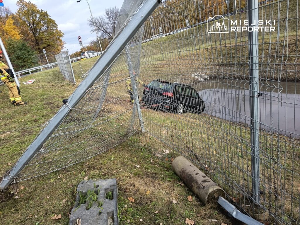 Czarny samochód utknął w rowie obok uszkodzonego ogrodzenia, a w tle widać strażaka w mundurze.