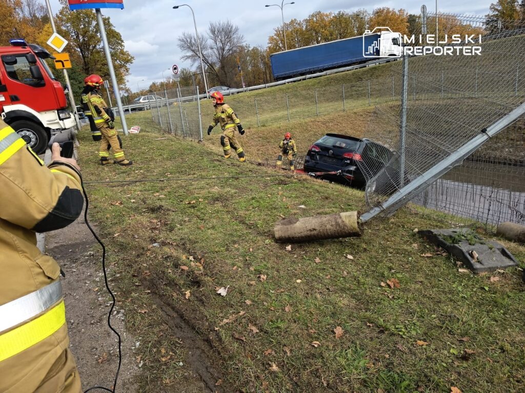 Strażacy w hełmach pracują nad wydobyciem samochodu z rowu, obok widoczny uszkodzony płot.