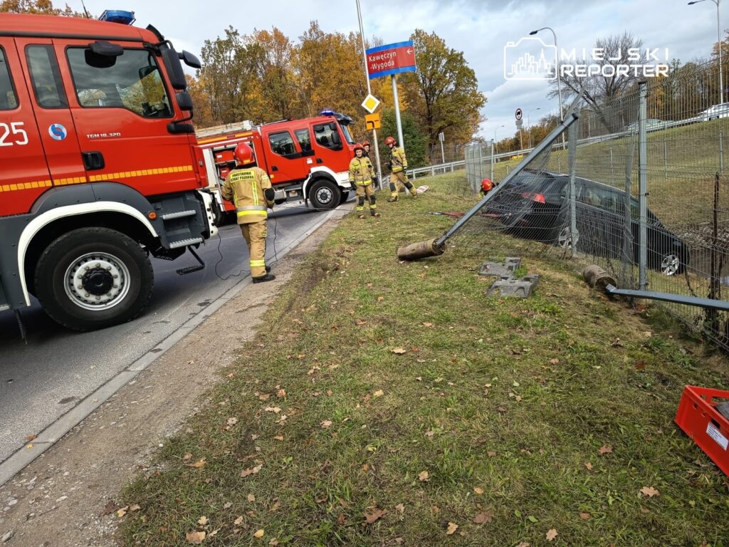 Strażacy w mundurach pracują przy wypadku drogowym, obok uszkodzonego samochodu i przewróconego słupa ogrodzeniowego.