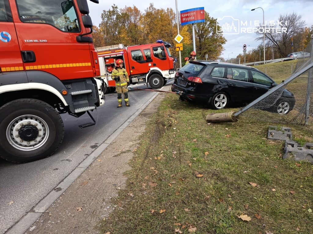 Strażak w mundurze kieruje akcją ratunkową przy przewróconym samochodzie na poboczu drogi, obok wozu strażackiego.