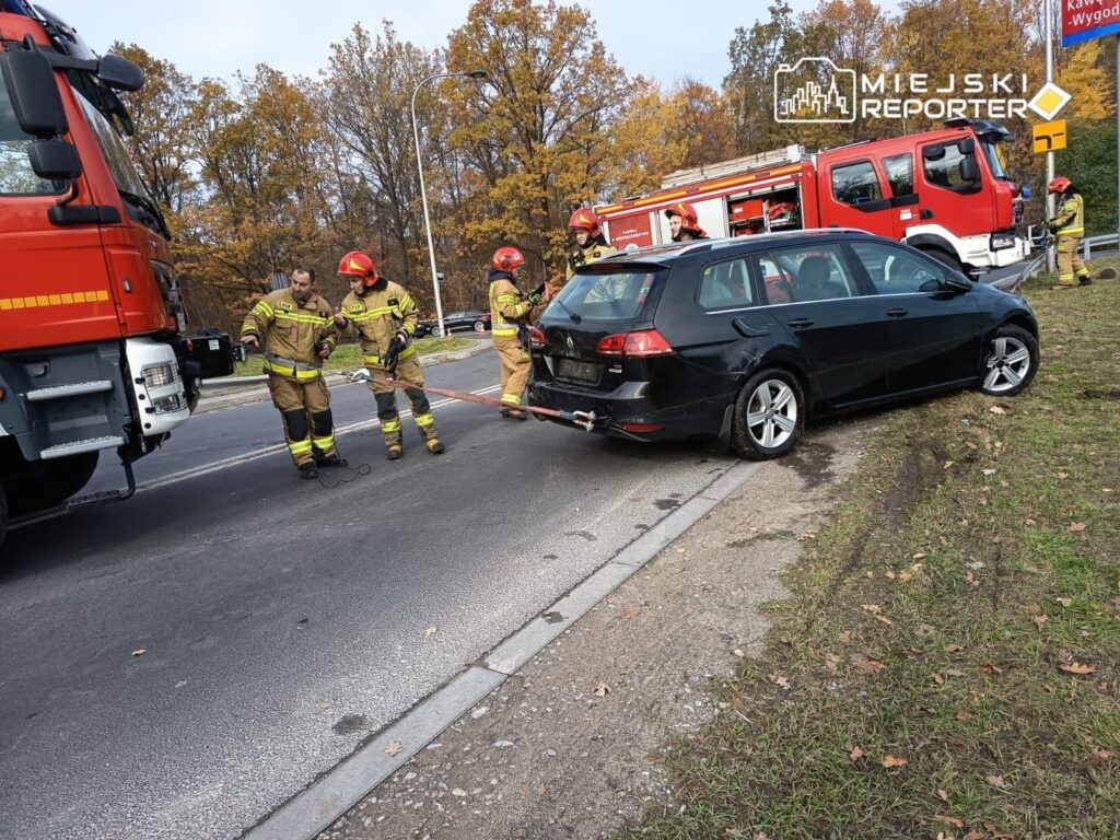Strażacy w mundurach zabezpieczają czarny samochód osobowy, który utknął na poboczu drogi.