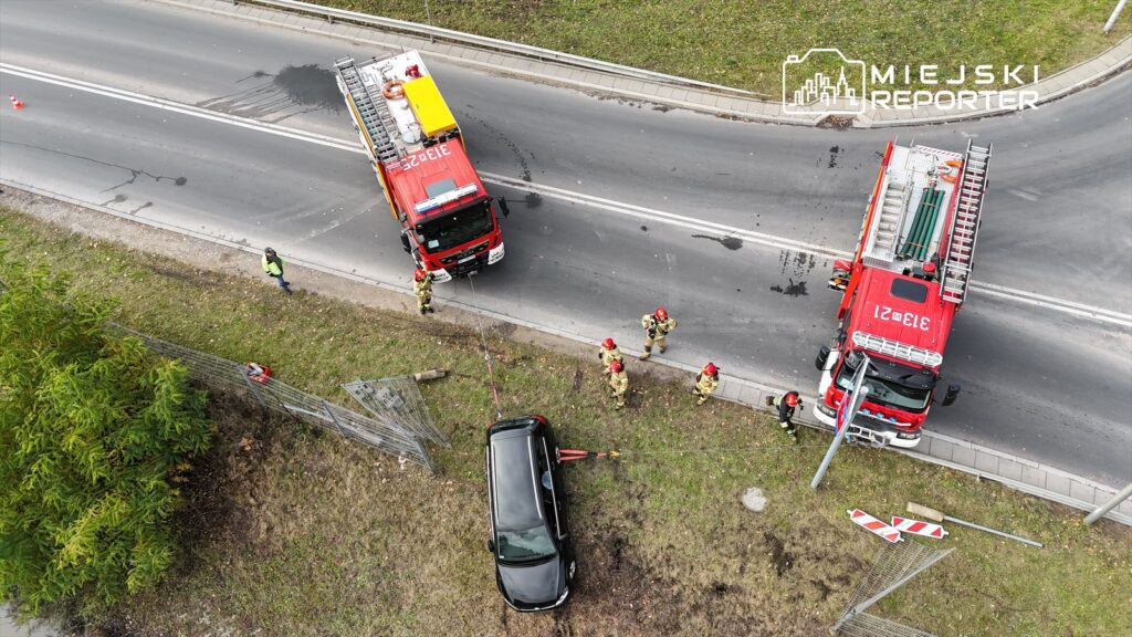 Strażacy w hełmach pracują przy wypadku drogowym, ciągnąc czarny samochód z pobocza na ruchliwej drodze.