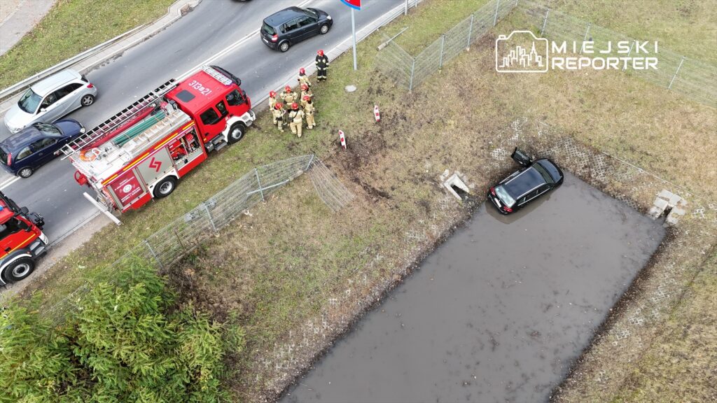 Czarny samochód utknął w wodzie obok drogi, a obok niego stoi grupa strażaków w mundurach, przygotowanych do akcji.