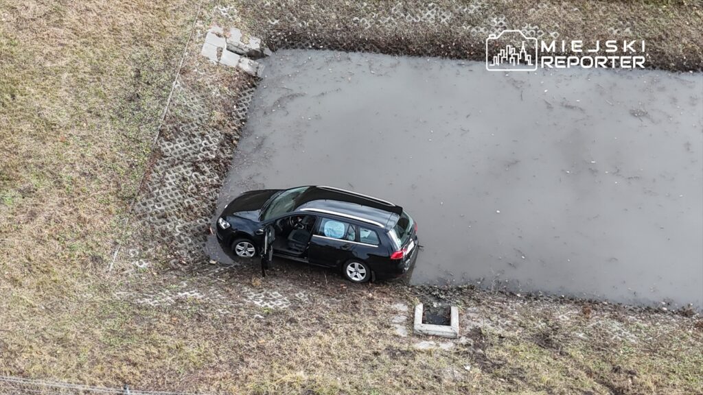 Czarny samochód osobowy zaparkowany na brzegu zarośniętego stawu, z otwartymi drzwiami po stronie kierowcy.