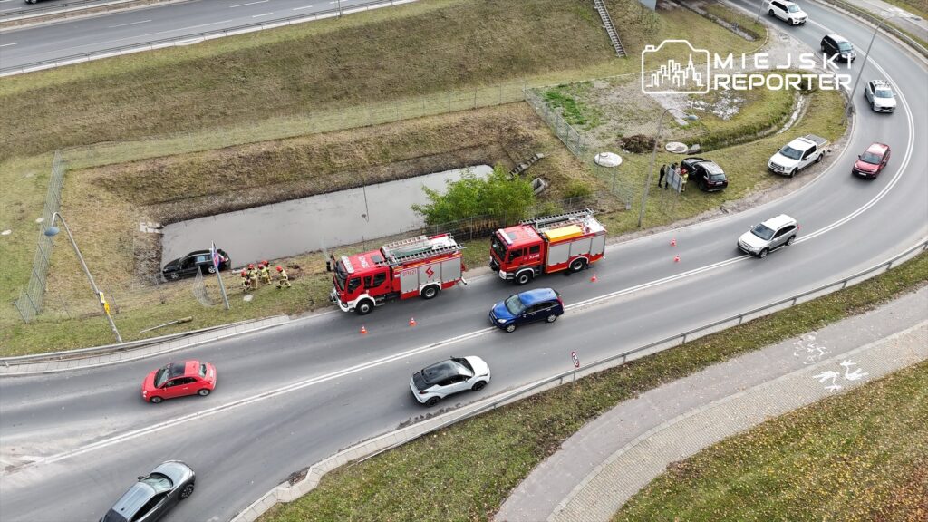 Strażacy w mundurach pracują przy czarnym samochodzie, który wpadł do rowu obok drogi, obok dwóch wozów strażackich.