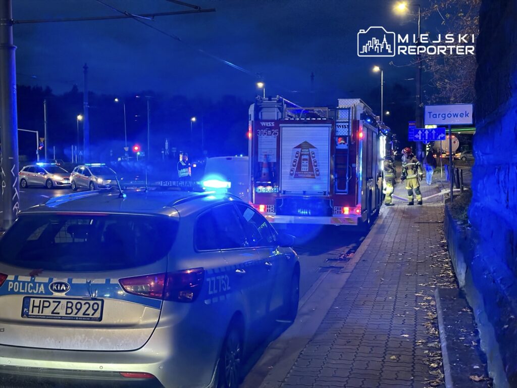 Policjant kierujący ruchem obok wozu strażackiego, w tle migające niebieskie światła radiowozów na ulicy.