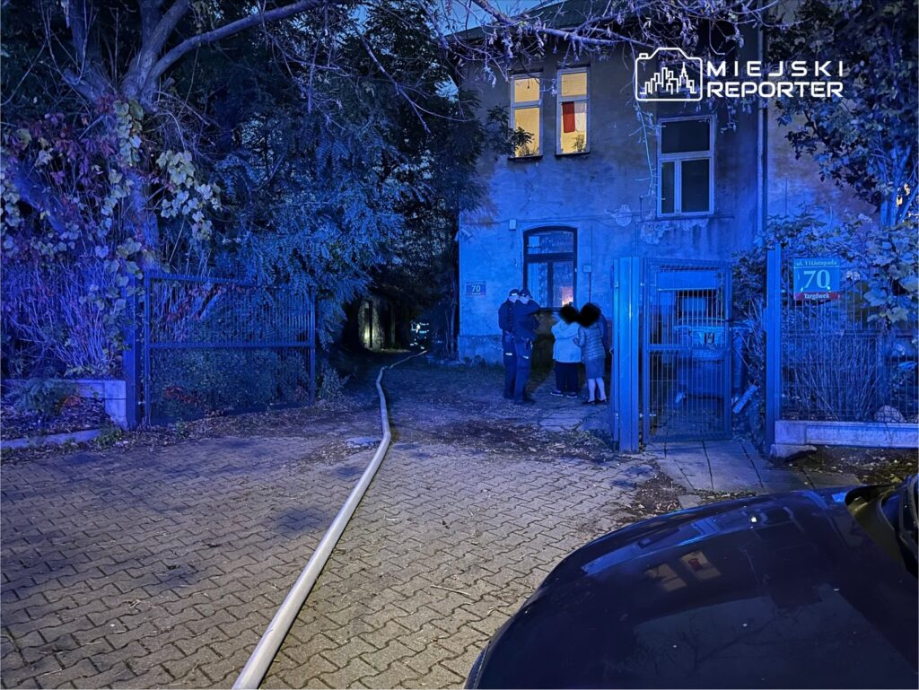 Grupa osób rozmawia przy wejściu do budynku, obok widoczna węża strażackiego na kostce brukowej.