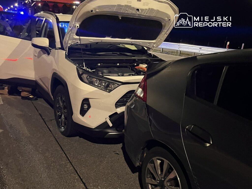 Dwa uszkodzone samochody na drodze, biały SUV z otwartą maską stoi obok ciemnego pojazdu po kolizji.