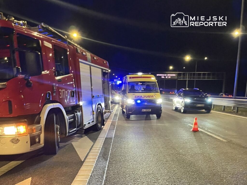 Na drodze nocą stoi wóz strażacki oraz karetka, a obok nich znajduje się czarny samochód osobowy. W tle widoczne są światła policyjne i znaki drogowe.