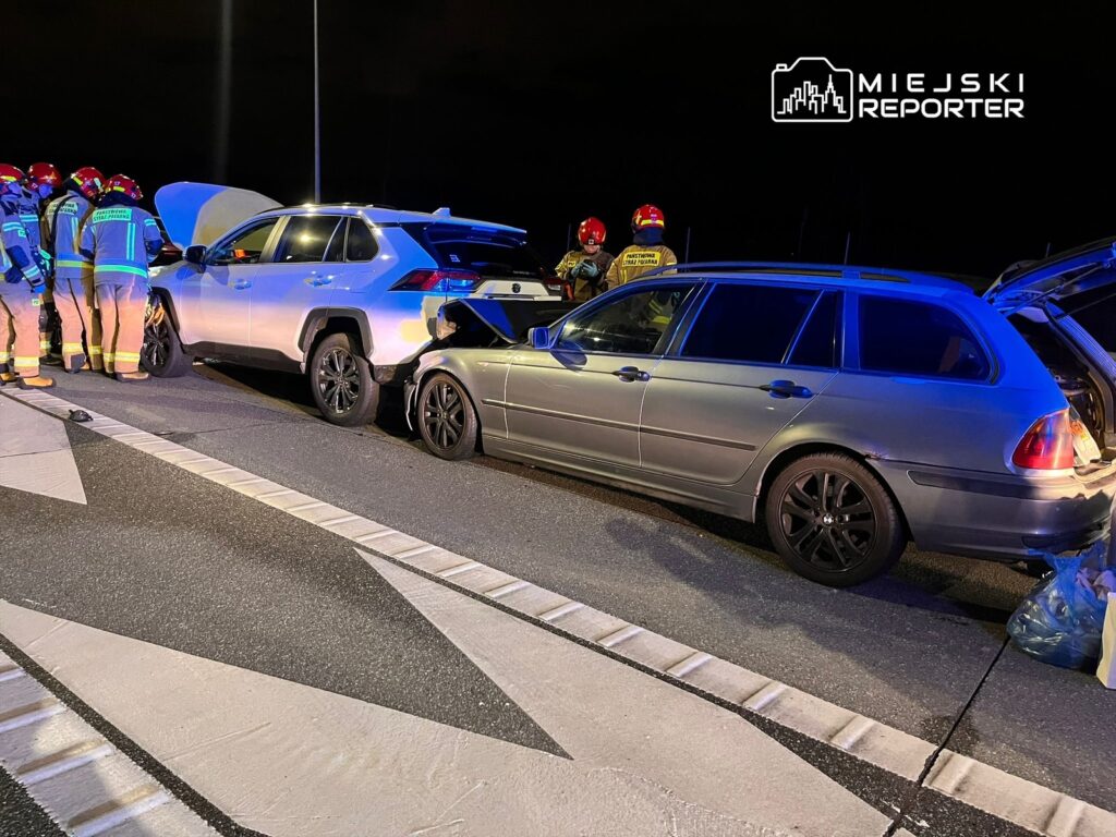 Grupa strażaków w mundurach bada uszkodzone pojazdy na drodze po wypadku, w nocy.