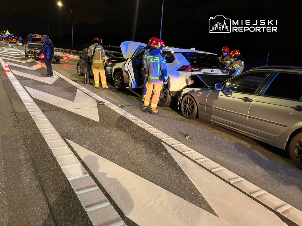 Grupa strażaków w mundurach i kaskach bada samochody po wypadku drogowym na autostradzie w nocy.