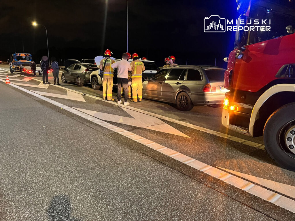 Grupa strażaków w kaskach rozmawia z kierowcami przy zderzeniu kilku samochodów na drodze w nocy.