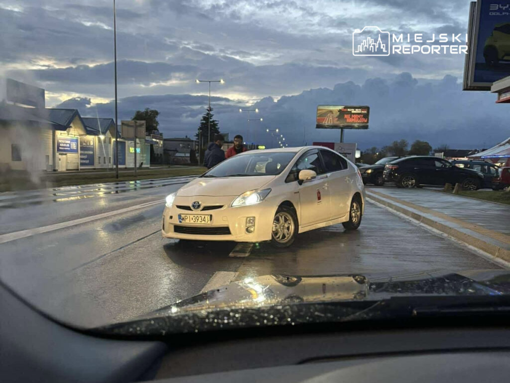 Biały samochód osobowy stoi na mokrej jezdni, obok dwie osoby rozmawiają, w tle widoczne budynki i billboard.