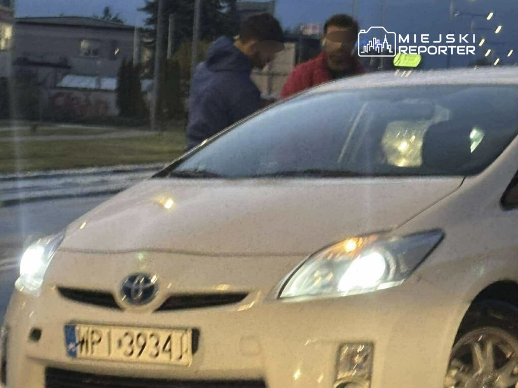 Dwaj mężczyźni rozmawiają obok białego samochodu osobowego na ulicy w deszczowy wieczór.