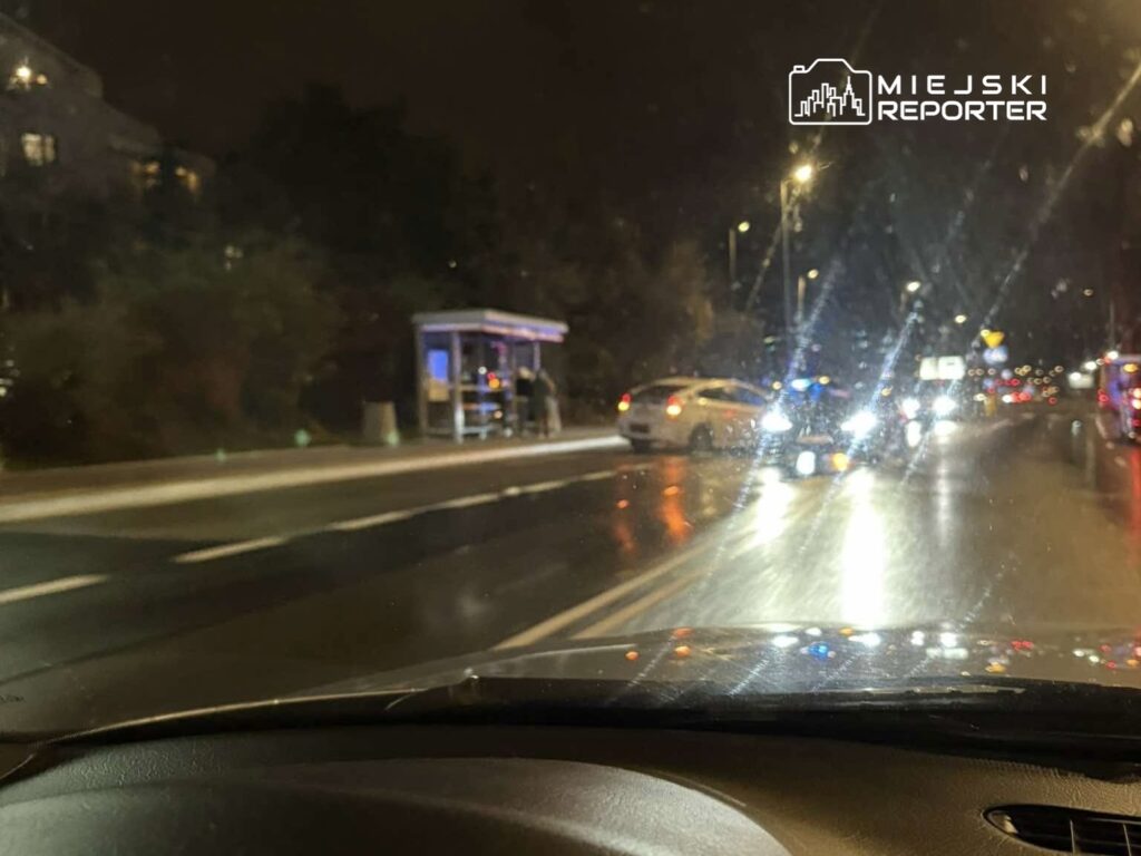 Osoby czekające na przystanku autobusowym w nocy, obok przejeżdżających samochodów i włączonych świateł policyjnych.
