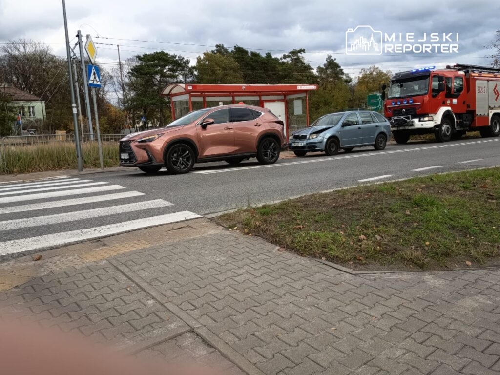 Luksusowy samochód osobowy w kolorze różowym zatrzymany przy przejściu dla pieszych obok niebieskiego auta i wozu strażackiego.