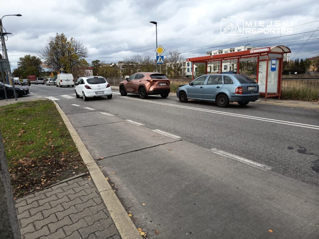 Ruch samochodowy na ulicy z przystankiem autobusowym po lewej stronie, w tle chmurne niebo.