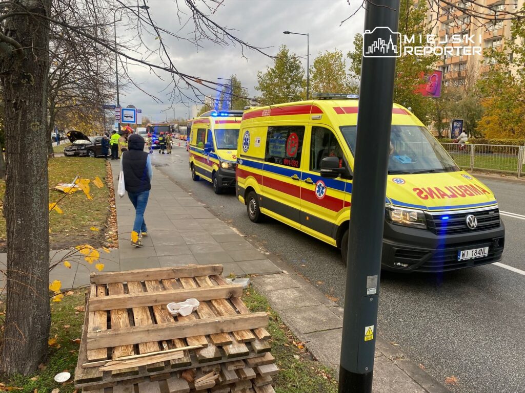Na chodniku obok drogi stoi mężczyzna w granatowej kamizelce, podczas gdy w tle pracują służby ratunkowe przy wypadku samochodowym.