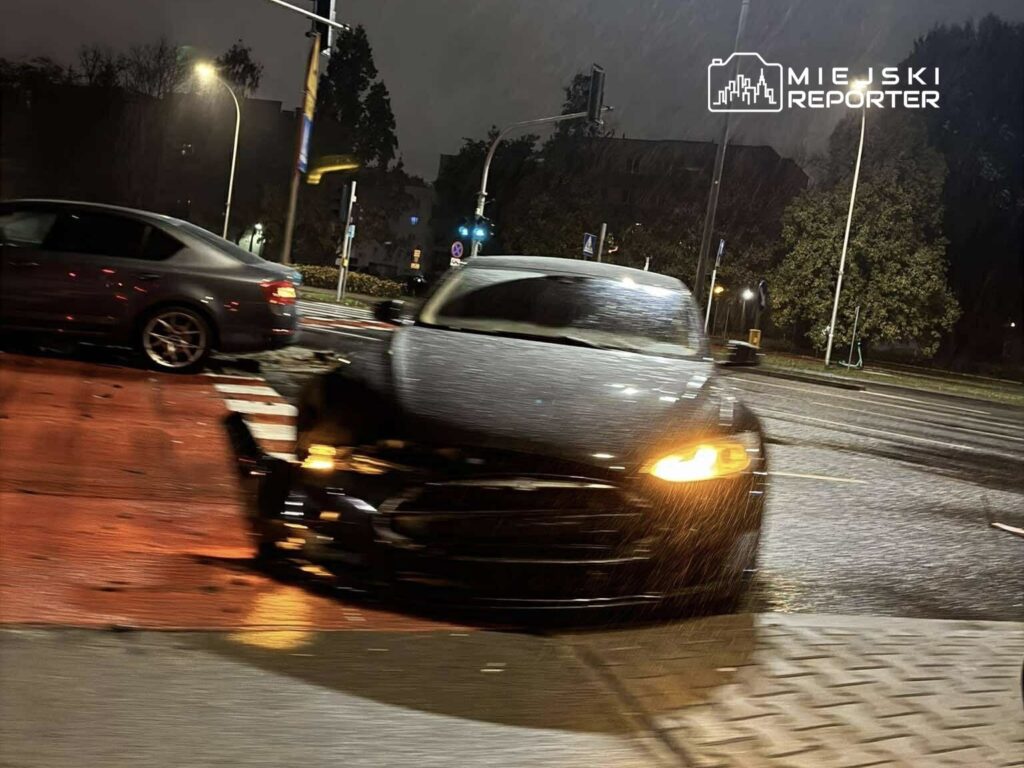 Czarny samochód osobowy z uszkodzoną przednią częścią stoi na skrzyżowaniu w deszczową noc.