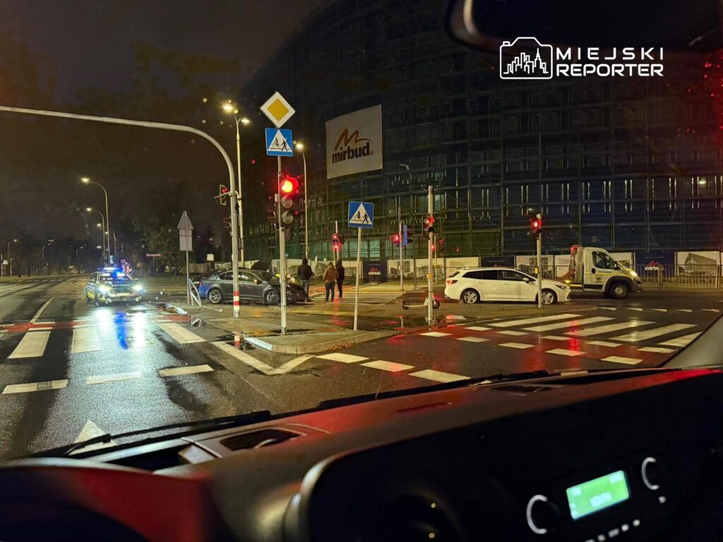 Policyjny radiowóz zatrzymany na skrzyżowaniu, obok uszkodzony samochód osobowy oraz kilku przechodniów w nocy.