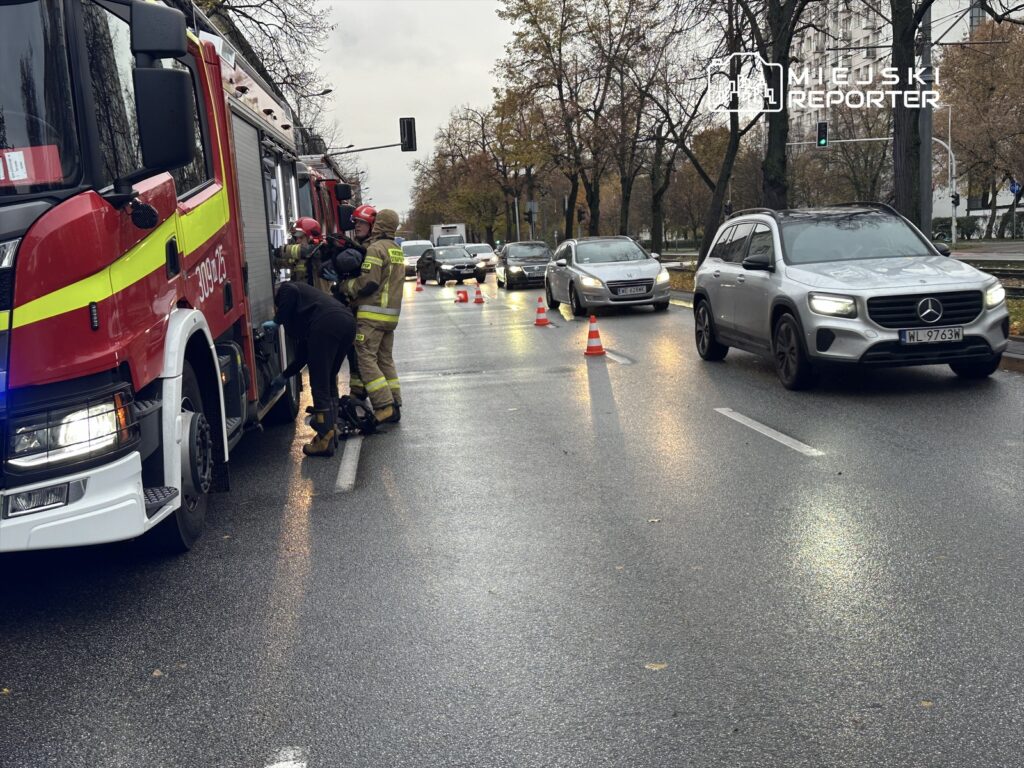 Strażacy w mundurach pracują przy wozie strażackim na mokrej jezdni, obok stoją samochody z ustawionymi pachołkami.