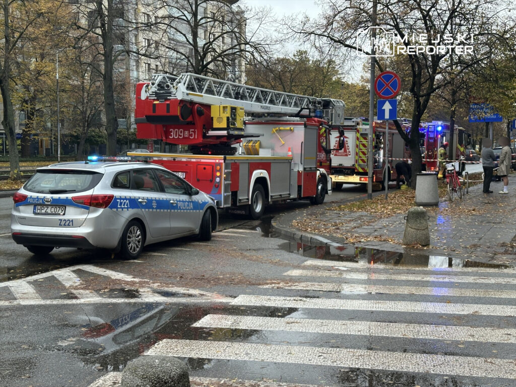 W tle widać strażacką drabinę, a przed nią zaparkowany radiowóz policji. Na chodniku stoją osoby w rozmowie.