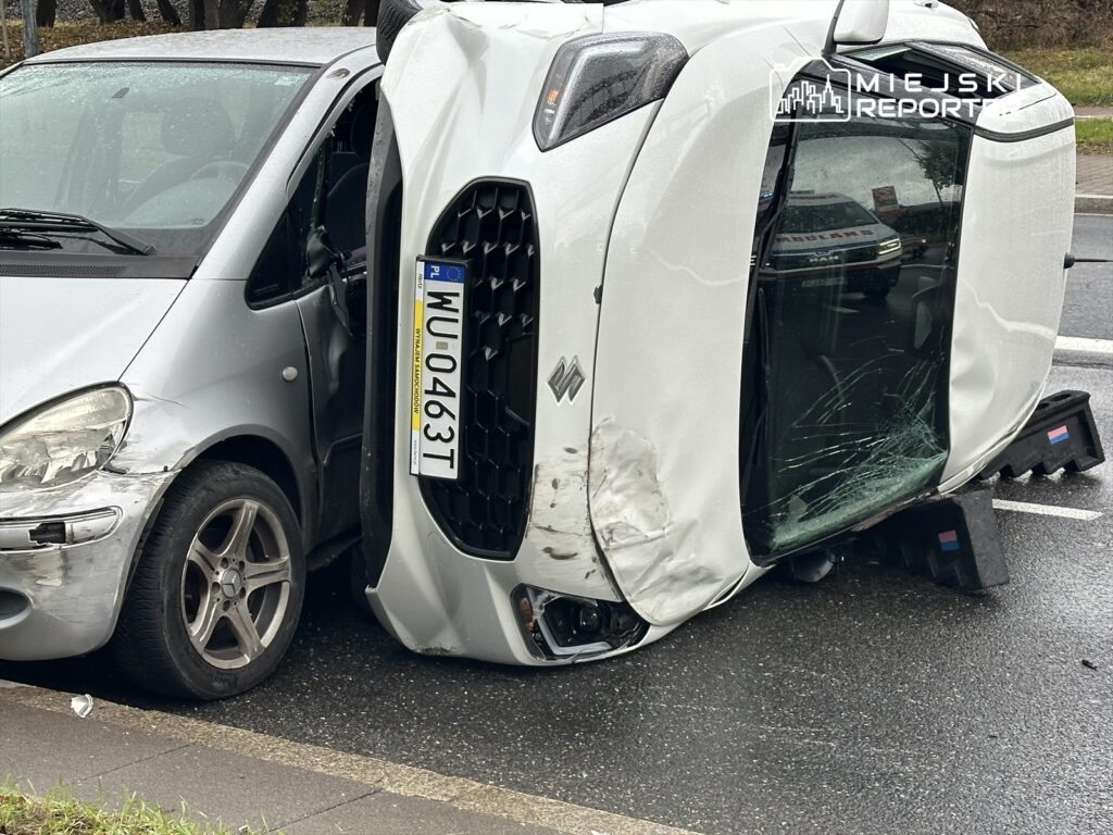 Dwa uszkodzone samochody osobowe, jeden przewrócony na bok, na deszczowej drodze.