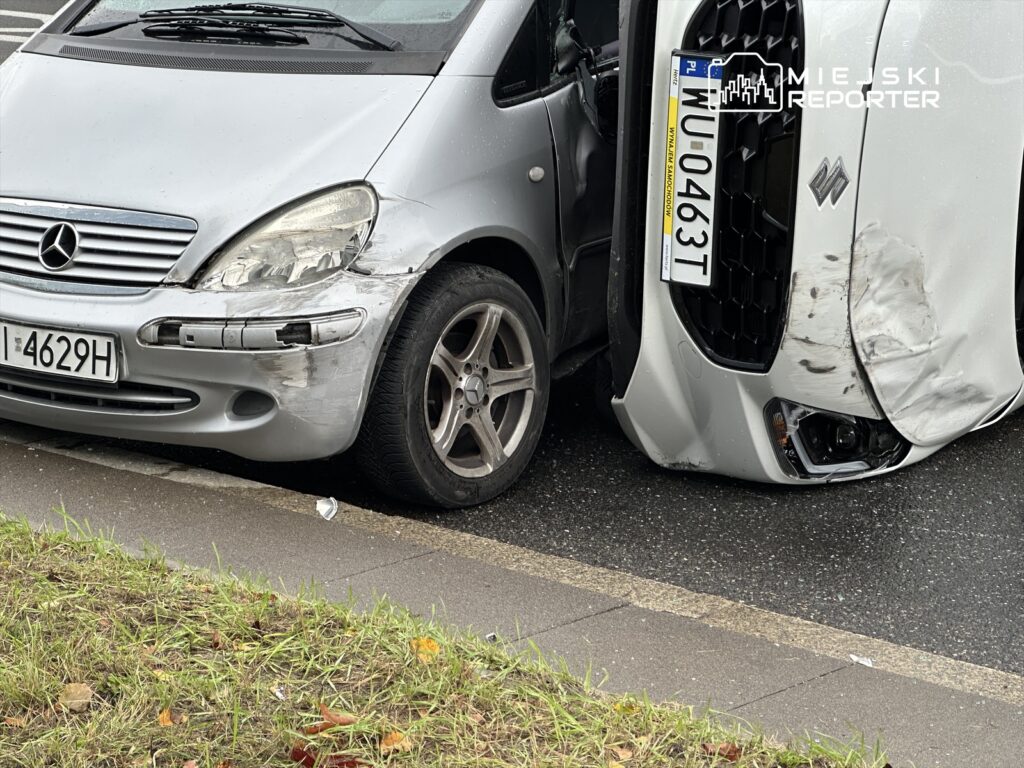 Dwa uszkodzone samochody osobowe stoją na poboczu drogi, jeden z nich leży na boku.