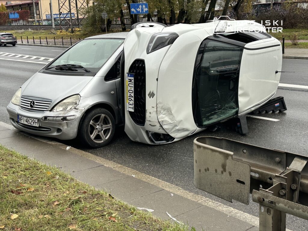 Dwa uszkodzone samochody na drodze, jeden z nich przewrócony na bok, obok leży metalowa barierka.