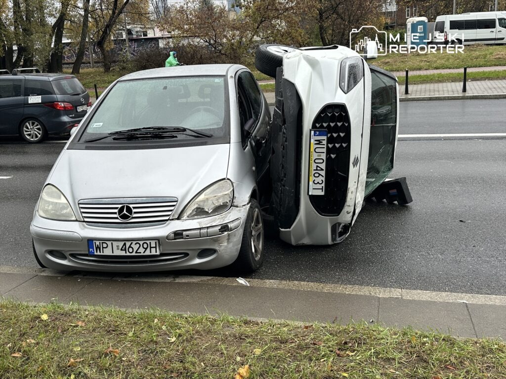 Złoty samochód osobowy leży na boku obok srebrnego vana na ruchliwej ulicy. W tle widać inne pojazdy oraz zieleń.